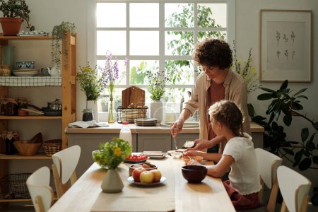 Photo for Young woman making sandwiches for youthful daughter and herself for breakfast while standing by kitchen table next to girl eating - Royalty Free Image