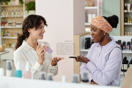 Foto de Joven asistente de tienda sonriente recomendando producto cosmético de mujer negra en una pequeña botella de plástico con pulverizador en el supermercado - Imagen libre de derechos