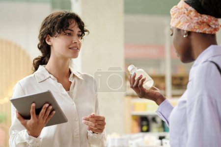 Photo for Young saleswoman with tablet looking at female customer with plastic bottle containing liquid self care product and consulting her - Royalty Free Image
