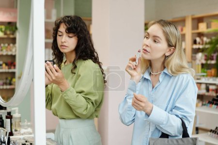 Téléchargez les photos : Jeune blonde femme test correcteur bâton sur son visage tout en se tenant devant le miroir dans le supermarché cosmétique - en image libre de droit