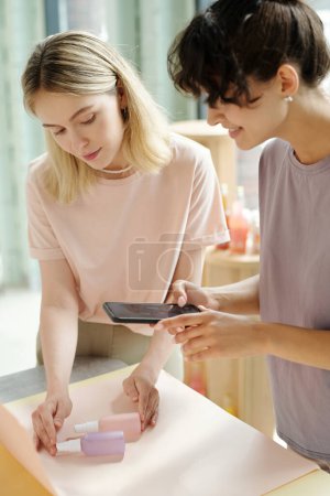 Photo for Young brunette woman with smartphone taking photo of beautycare products while her colleague putting them on white paper - Royalty Free Image