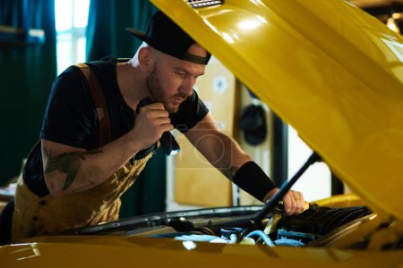Téléchargez les photos : Jeune mécanicien d'atelier de réparation automobile vérifiant le moteur de l'automobile jaune tout en dirigeant flashligh sur son moteur - en image libre de droit