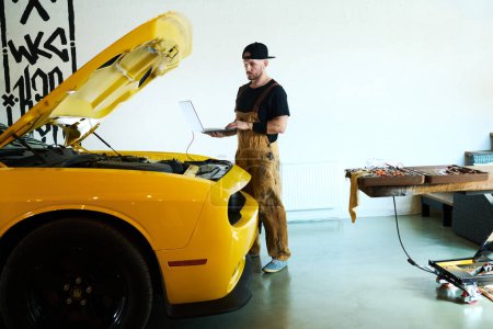 Téléchargez les photos : Jeune mécanicien en tenue de travail portable pendant les diagnostics techniques du moteur et d'autres détails de voiture électrique de couleur jaune - en image libre de droit