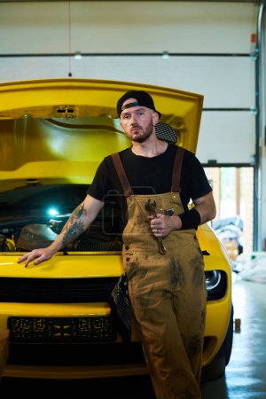 Foto de Joven trabajador serio del servicio de mantenimiento de automóviles de pie por automóvil de color amarillo con capucha abierta y mirando a la cámara - Imagen libre de derechos