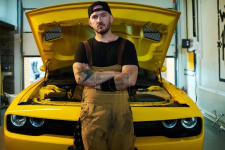Photo for Young serious workman in overalls, blackc t-shirt and baseball cap keeping his arms crossed on chest while standing by car - Royalty Free Image