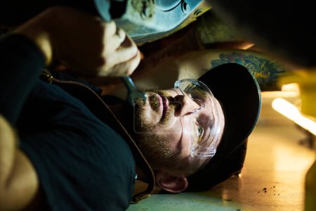 Photo for Close-up of young repairman in protective eyeglasses and workwear lying under car while cheking its condition and fixing details with wrench - Royalty Free Image