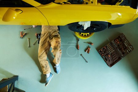 Téléchargez les photos : Vue ci-dessus du réparateur en salopette et en chewing-gums couchés sous une voiture jaune sur le sol de l'atelier ou du garage et fixation des détails avec des outils de travail - en image libre de droit
