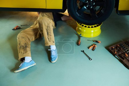 Téléchargez les photos : Jambes de jeune réparateur ou technicien en salopette couchées sous une voiture électrique jaune sur le sol de l'atelier ou garae et le réparer - en image libre de droit