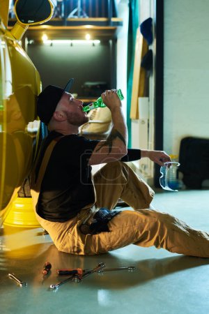 Photo for Young tired mechanic in workwear drinking beer from bottle while sitting on the floor by yellow car after repairment or technical checkup - Royalty Free Image