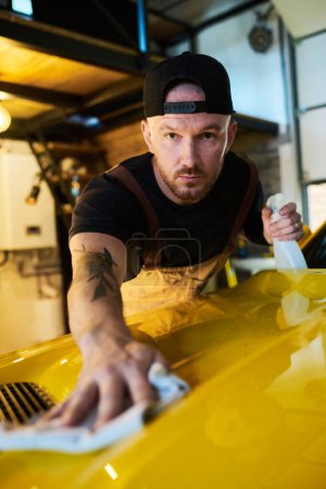 Foto de Joven trabajador masculino del servicio de mantenimiento con botella de detergente cubierta de la campana de limpieza de automóvil amarillo después de los trabajos de reparación - Imagen libre de derechos