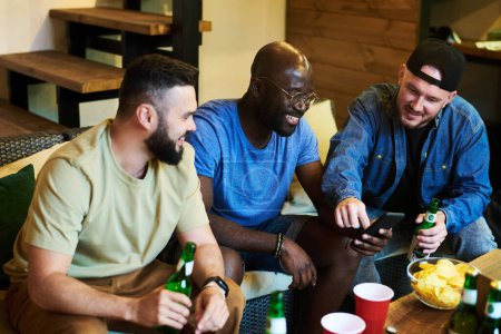 Téléchargez les photos : Copains joyeux discutant de photos dans le smartphone du gars afro-américain assis à table avec des bouteilles de bière et des collations - en image libre de droit