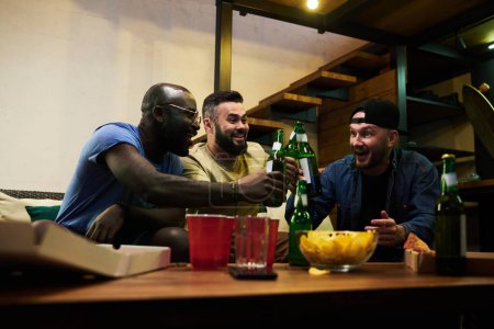 Junge fröhliche interrassische Kumpels und Fußballfans, die mit Bierflaschen über einen Tisch mit Tassen und Snacks klirren, während sie den Sieg feiern
