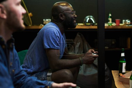 Téléchargez les photos : Joyeux jeune homme afro-américain en t-shirt bleu jouant au jeu vidéo tout en étant assis sur le canapé et en appuyant sur les boutons sur la manette de jeu - en image libre de droit