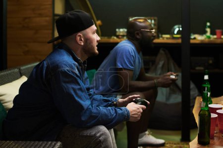 Foto de Hombre tenso en ropa de casualwear presionando botones en el joystick mientras juega videojuego con su amigo mientras ambos están sentados frente a la mesa con cerveza - Imagen libre de derechos