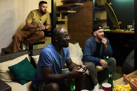 Téléchargez les photos : Deux jeunes hommes avec de la bière regardant l'écran de télévision et regardant le jeu vidéo tandis que le gars noir avec joystick assis à proximité et en appuyant sur des boutons - en image libre de droit