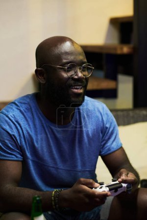 Téléchargez les photos : Jeune homme noir souriant en lunettes et t-shirt bleu appuyant sur les boutons sur le contrôleur tout en jouant à un jeu vidéo devant l'écran de télévision - en image libre de droit