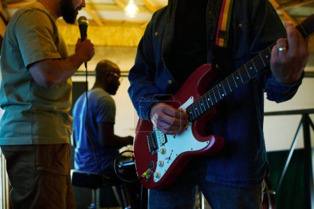 Foto de Primer plano del joven en chaqueta de mezclilla y jeans azules tocando guitarra eléctrica contra cantante con micrófono y batería - Imagen libre de derechos