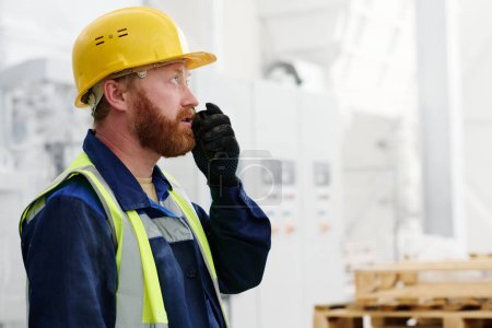 Téléchargez les photos : Travailleur masculin barbu en uniforme, casquette et gants noirs utilisant un talkie-walkie tout en signalant le processus de production au gestionnaire ou à l'employeur - en image libre de droit