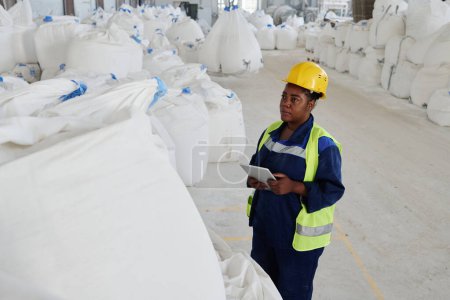 Photo for Young serious black woman in uniform and safety helmet using tablet while standing in front of huge white sack with raw materials - Royalty Free Image