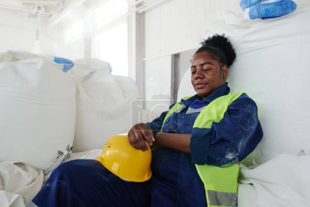 Téléchargez les photos : Ingénieur femelle fatiguée en uniforme tenant casque de sécurité tout en étant assise contre un énorme sac blanc dans l'entrepôt et la sieste - en image libre de droit