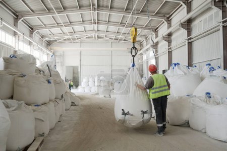 Photo for Rear view of young male worker of factory in uniform and hardhat moving along rows of huge heavy sacks with loose raw materials - Royalty Free Image