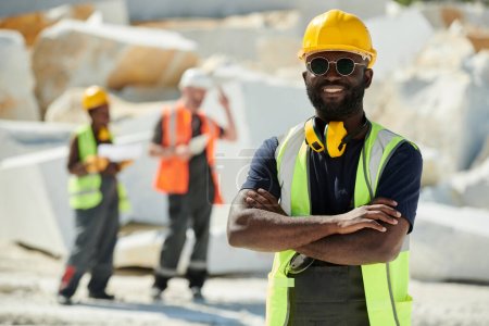 Photo for Young successful African American male worker of marble quarry with his arms crossed on chest standing in front of camera against colleagues - Royalty Free Image