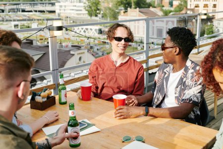 Photo for Young cheerful interracial men and their girlfriends in stylish casualwear chatting by table in rooftop cafe and having beer - Royalty Free Image