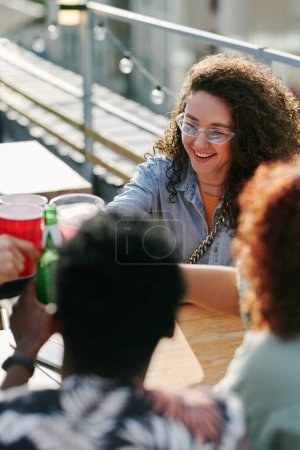 Photo for Young cheerful woman in eyeglasses and casualwear toasting with her friends gathered by table in outdoor cafe on summer weekend - Royalty Free Image