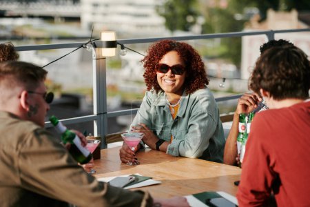 Téléchargez les photos : Jeune femme noire souriante avec cocktail rose en verre martini assis à table devant ses amis profitant du rassemblement dans un café en plein air - en image libre de droit