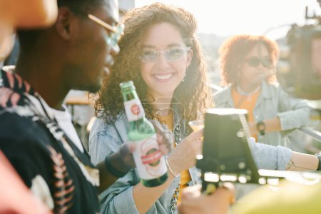 Téléchargez les photos : Fille heureuse avec cocktail parlant à son petit ami afro-américain avec bouteille de bière tout en bavardant dans un café en plein air le week-end d'été - en image libre de droit