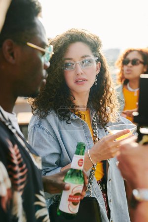 Foto de Mujer joven con cóctel mirando al hombre afroamericano con botella de cerveza mientras ambos disfrutan de la fiesta en la cafetería al aire libre - Imagen libre de derechos