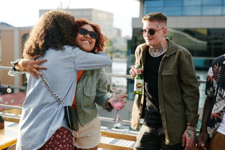 Photo for Happy young black woman with cocktail in martini glass giving hug to friend while standing against guy with bottle of beer - Royalty Free Image