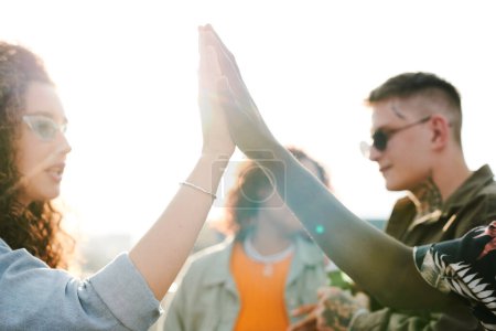 Photo for Young intercultural dates giving each other high five against their friends having chat during outdoor party in rooftop cafe or restaurant - Royalty Free Image