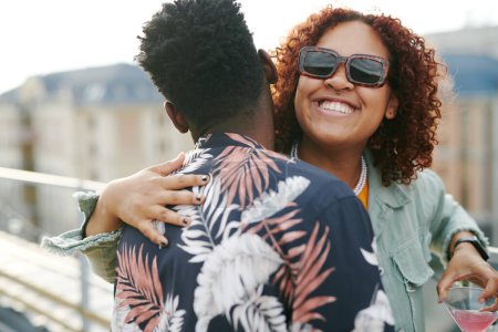 Téléchargez les photos : Jeune femme noire joyeuse dans des lunettes de soleil donnant câlin à l'homme afro-américain tout en profitant de la fête sur le toit - en image libre de droit