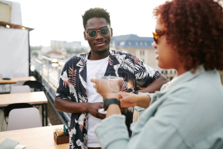 Photo for African American guy in sunglasses standing in front of his girlfriend and chatting to her while both having drinks in rooftop cafe - Royalty Free Image