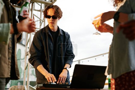 Téléchargez les photos : Jeune DJ masculin en tenue décontractée et lunettes de soleil faire de la musique pour ses amis avec de la bière et des cocktails profiter de la fête sur le toit - en image libre de droit