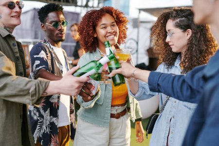 Foto de Grupo de felices jóvenes amigos interculturales en ropa casual tostadas con cerveza y cócteles y disfrutar de la fiesta en la azotea el fin de semana - Imagen libre de derechos
