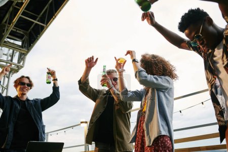 Téléchargez les photos : Groupe de jeunes amis extatiques avec des bouteilles de bière dansant à la fête sur le toit dans un café en plein air tout en se tenant les bras levés - en image libre de droit