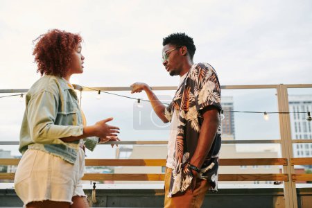 Téléchargez les photos : Jeune femme noire en tenue décontractée expliquant quelque chose à son petit ami tout en se tenant devant lui sur la terrasse du café sur le toit - en image libre de droit