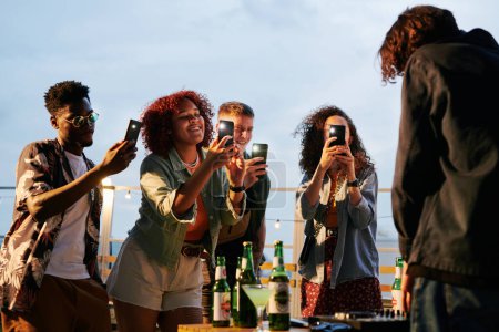 Téléchargez les photos : Deux jeunes couples interculturels heureux avec smartphones prenant des photos de DJ à la fête sur le toit tout en se tenant devant lui - en image libre de droit