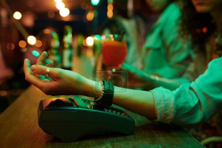 Téléchargez les photos : Main de jeune cliente avec smartwatch au poignet sur l'écran du terminal de paiement debout sur le comptoir dans le bar ou le café - en image libre de droit