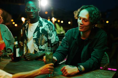 Téléchargez les photos : Heureux jeune homme achetant mojito cocktail tout en se tenant au comptoir dans le bar contre le couple afro-américain profiter de soirée - en image libre de droit