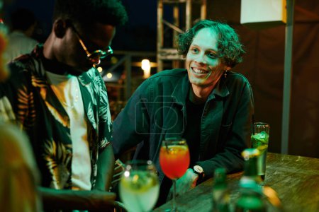 Téléchargez les photos : Jeune homme heureux en tenue décontractée noire regardant un de ses amis pendant le chat tout en étant assis au comptoir avec des cocktails commandés dans le bar - en image libre de droit