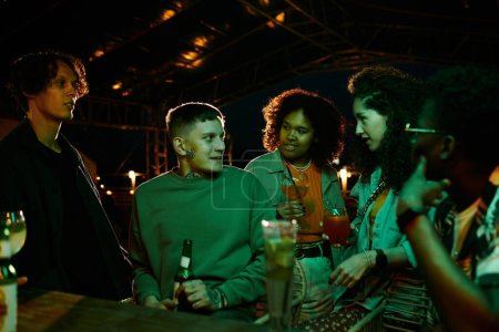 Téléchargez les photos : Jeunes filles interculturelles avec des cocktails et des gars à l'écoute du jeune homme avec bouteille de bière tout en profitant de la fête dans une boîte de nuit - en image libre de droit