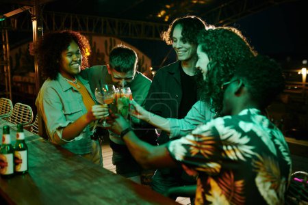 Foto de Grupo de alegres amigos interculturales tintinean con copas de cócteles mientras están de pie junto al mostrador del bar y disfrutan de la fiesta en el club nocturno - Imagen libre de derechos