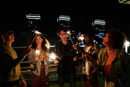 Foto de Grupo de amigos multiculturales jóvenes felices con luces de bengala brillantes disfrutando de la fiesta en la cafetería de la azotea en la noche y divirtiéndose - Imagen libre de derechos