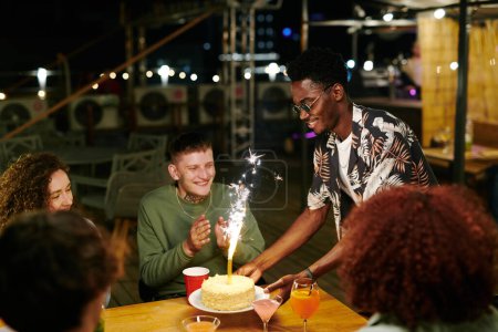 Foto de Feliz joven afroamericano poniendo plato con sabroso pastel con espumoso en la mesa festiva donde sus amigos se reunieron para la fiesta - Imagen libre de derechos