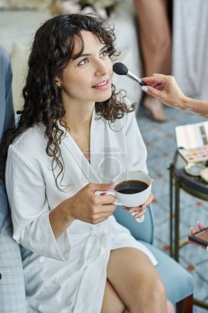 Téléchargez les photos : Jeune femme en peignoir blanc ayant expresso tout en regardant le visage artiste pendant la procédure de maquillage le matin du mariage - en image libre de droit