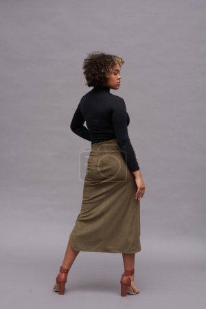 Téléchargez les photos : Vue arrière de la jeune femme afro-américaine élégante à col roulé et jupe longue debout devant la caméra en studio et regardant de côté - en image libre de droit