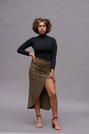 Téléchargez les photos : Jeune femme afro-américaine bien habillée en col roulé et jupe longue regardant la caméra tout en se tenant debout avec la main droite sur sa taille - en image libre de droit